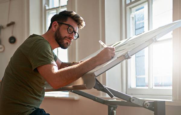 Man in glasses sitting sketching at a desk.