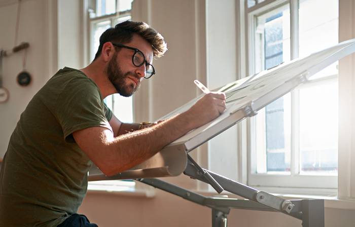 Man in glasses sitting sketching at a desk. 